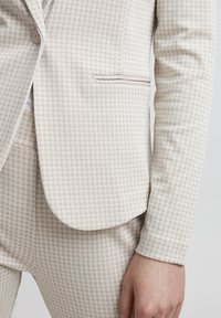 Svetle béžový blejzr s houndstooth vzorem, s klopovým límcem, zapínáním na jeden knoflík a hladkou texturou.