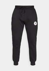 Black cotton sweatpants with a drawstring waist, tapered legs, and a circular logo patch on the left thigh. Soft texture, elastic cuffs.