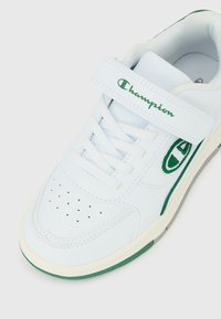 Λευκό αθλητικό παπούτσι Champion με πράσινο λογότυπο, κορδόνια στο μπροστινό μέρος και λουράκι με βέλκρο, ορατό από πάνω και με γωνία για να φαίνονται οι πλευρικές λεπτομέρειες.