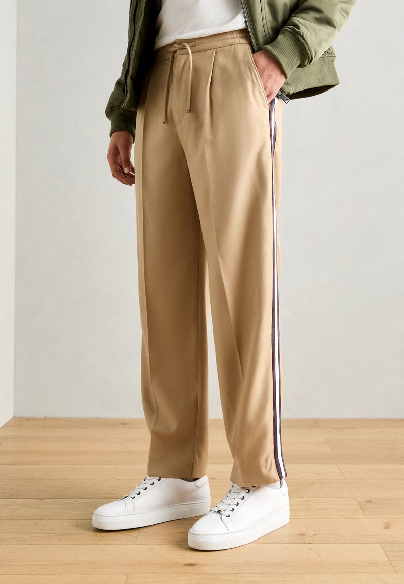 Beige bukser med en afslappet pasform, elastisk talje, frontfolder og kontrasterende sidelister; parret med hvide sneakers på et trægulv.