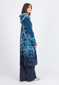 Lange blauwe gebreide jas met een capuchon, ingewikkelde patronen in verschillende tinten blauw en een zijsluiting met knopen. Gedragen over wijde spijkerbroek.