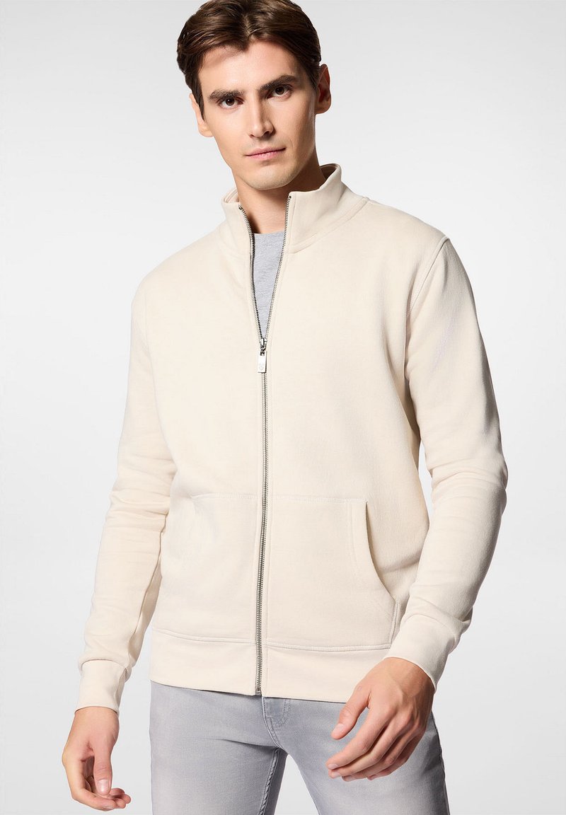 Beżowa rozpinana bluza z miękkiego materiału, z wysokim kołnierzem, dwoma przednimi kieszeniami i ściągaczami na rękawach. Nosi się ją na jasnoszarej koszuli.