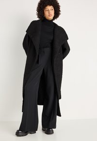 Lungo cappotto nero dalla texture marcata con ampio colletto, indossato sopra un dolcevita nero. Pantaloni neri a gamba larga su misura e scarpe nere chunky.