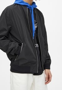 Veste bomber noire à finition lisse et brillante, avec une fermeture éclair à l'avant, des poignets côtelés et un sweat à capuche bleu porté en dessous.