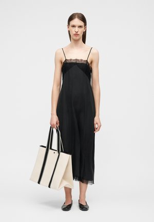 Γυναίκα που φοράει μαύρο φόρεμα slip με δαντελένιο τελείωμα και μαύρα φλατ παπούτσια, κρατώντας μια μεγάλη κρεμ τσάντα ώμου με μαύρες κάθετες ρίγες.