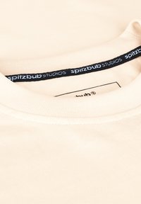 Helles beiges T-Shirt mit einem gerippten Rundhalsausschnitt, ausgestattet mit einem schwarzen Etikett mit "spitzbub STUDIOS" in weißem Text im Kragen.