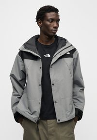 Γκρι αδιάβροχο μπουφάν με κουκούλα, διαθέτει μαύρες πινελιές, κλείσιμο με φερμουάρ και κουμπιά με πατιλέτα. Φοριέται πάνω από μαύρο μπλουζάκι.