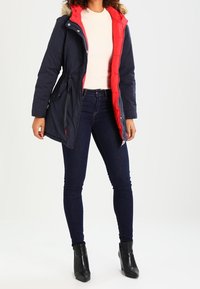 Námořnická a červená prošívaná bunda s kapucí lemovanou kožešinou, zipem vpředu a nastavitelným pasem. V kombinaci s tmavými džíny a černými kotníkovými botami.