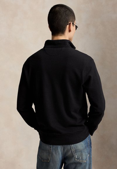 Hombre con una camisa negra de manga larga y pantalones vaqueros azules, de espaldas sobre un fondo beige liso.