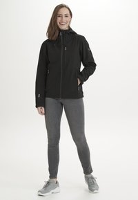Svart softshell-jacka med huva, framdragkedja, sidofickor och elastiska ärmslut, i kombination med grå jeans och grå sneakers.
