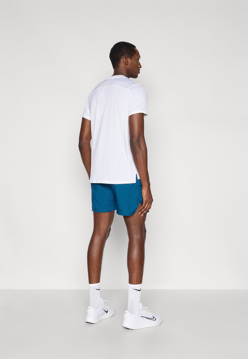 Vit pikétröja med korta ärmar, blå shorts, vita sneakers och vita strumpor, sedd bakifrån mot en ljusgrå bakgrund.