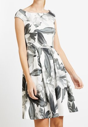 Kvinde iført hvid knælængde kjole med stort gråt blomsterprint, kapærmer og tætsiddende talje.