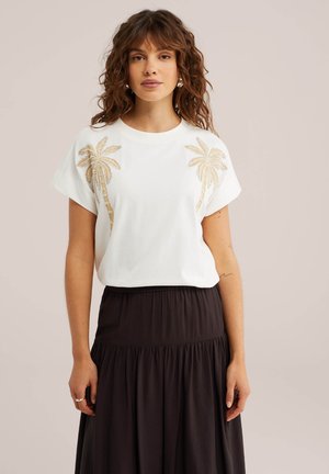 Donna che indossa una t-shirt bianca con disegni di palme dorate e gonna nera plissettata a vita alta, in piedi su uno sfondo semplice.