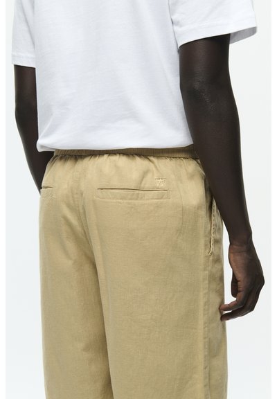 Beige Leinenhosen mit elastischem Bund, zwei Gesäßtaschen und einem kleinen gestickten Logo. Weißes T-Shirt mit kurzen Ärmeln.