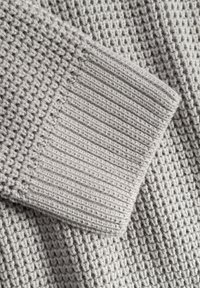 Nahaufnahme eines hellgrauen gestrickten Pullover-Stoffes, der den gerippten Bund und das strukturierte Stitch-Muster zeigt.