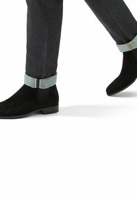 Botas de tobillo de ante negro con un acabado suave, contrastadas por pantalones de mezclilla con un dobladillo enrollado en un tono más claro y detalles de costura sutiles.