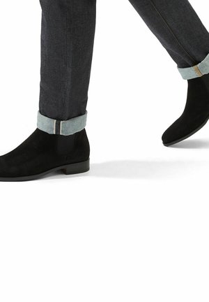 Botas de tobillo de ante negro con un acabado suave, contrastadas por pantalones de mezclilla con un dobladillo enrollado en un tono más claro y detalles de costura sutiles.