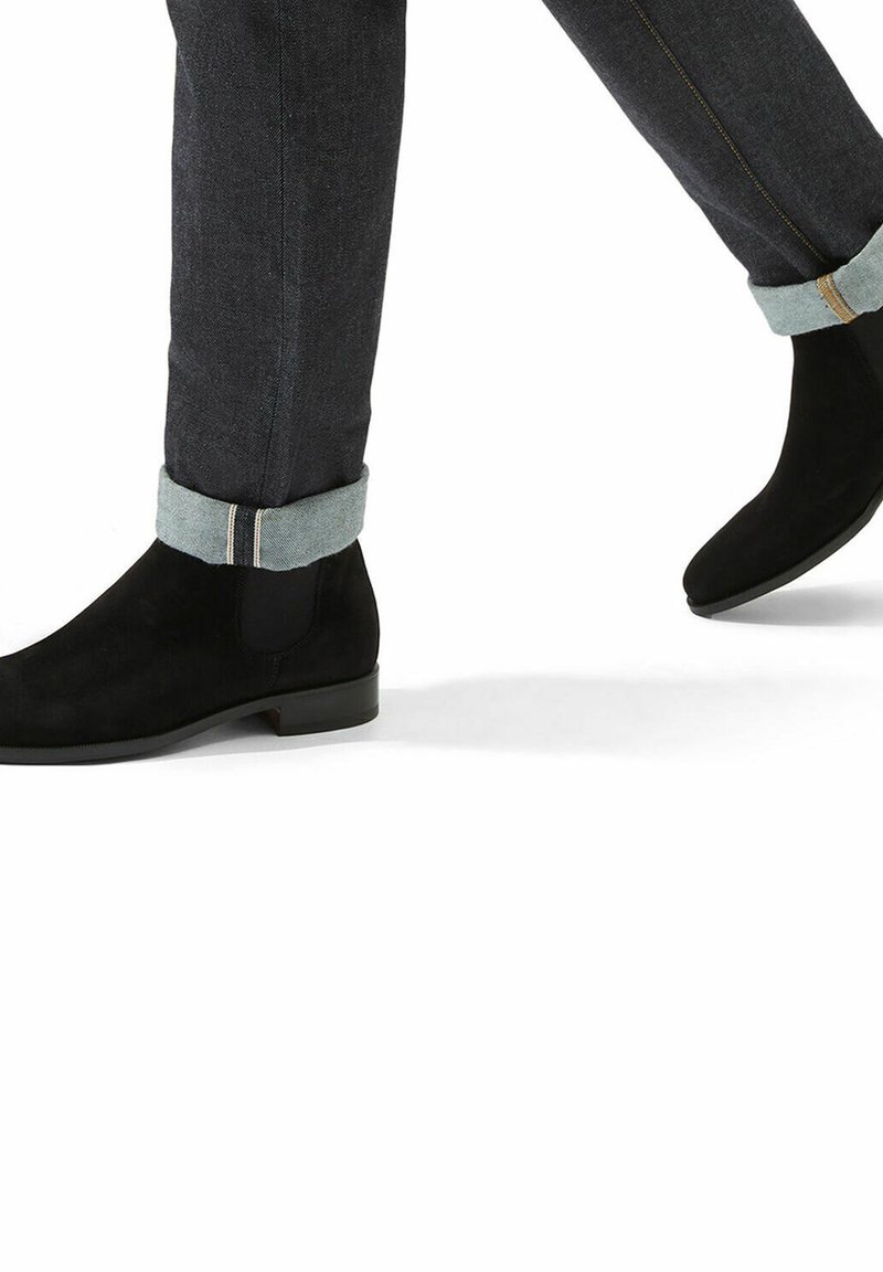 Botas de tobillo de ante negro con un acabado suave, contrastadas por pantalones de mezclilla con un dobladillo enrollado en un tono más claro y detalles de costura sutiles.