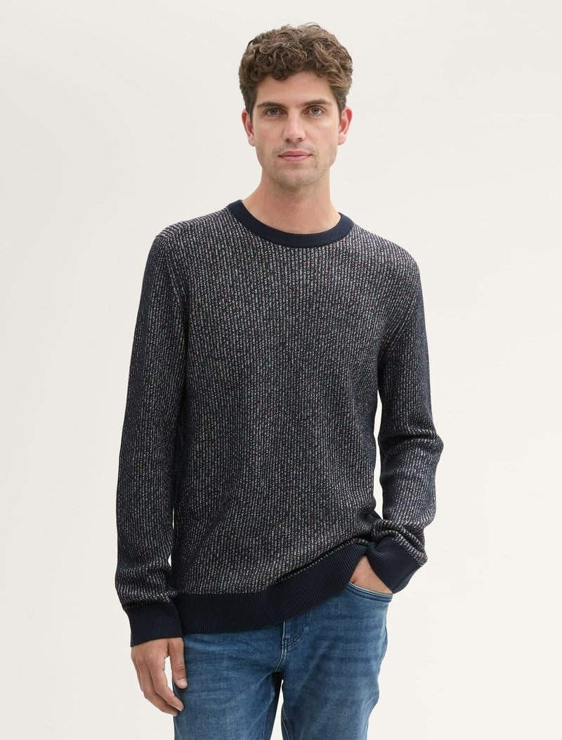 Maglione lavorato a maglia di colore blu scuro con motivo a strisce texturizzate; collo rotondo, polsini e orlo aderenti. Abbinato a jeans azzurri.