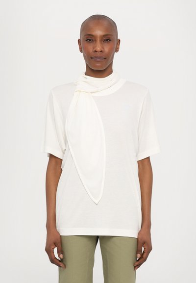 Weißes kurzärmliges T-Shirt mit hohem Kragen, das mit einem leichten weißen Schal um den Hals drapiert ist. Olivgrüne Hose vervollständigt den Look.