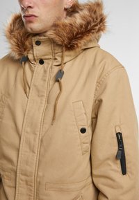 Beige parka med hette med fuskepels, glidelåslomme på siden, justerbare snorer og knappelukking. Glatt stoffstruktur.