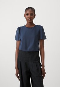 Mörkblå kortärmad blus med en slät yta och plisserade detaljer, i kombination med högt midjebyxor i svart med texturerad tyg.