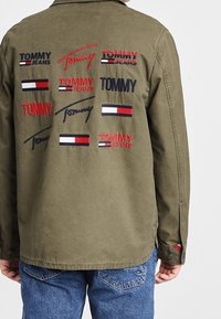 Olive green shirt with embroidered "TOMMY" logos in red and blue, featuring a button-down collar and long sleeves, paired with blue jeans.