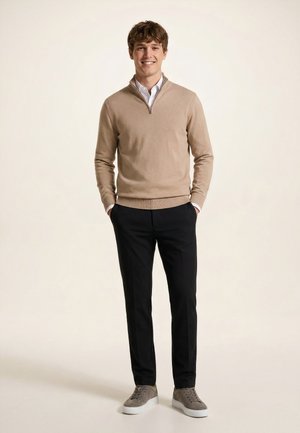 Mladý muž sa usmieva, nosí béžový sveter na štvrťový zips, bielu košeľu, čierne nohavice a sivé tenisky, stojí s rukami v vreckách.