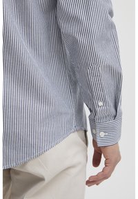 Camicia a righe in tessuto di cotone blu e bianco, con polsini abbottonati, yoke appuntito e orlo curvo. Texture liscia e vestibilità su misura.