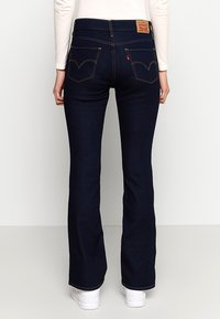 Breddet mørkeblå denimjeans med kontrasterende gul syning og to baglommer. Lædermærke på taljen. Glat tekstur.