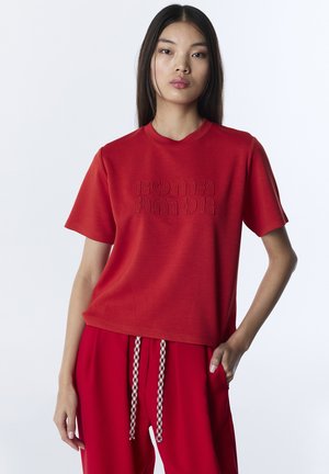 Mujer joven con cabello largo y oscuro que lleva una camiseta roja de manga corta con texto en relieve y pantalones rojos a juego con cordones estampados.