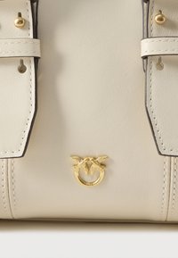 Bolsa de couro creme com detalhes em dourado, costura superior e um emblema único com duas aves pousadas em um círculo.