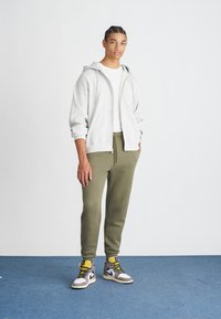 Sweatshirt cinza com fecho, t-shirt branca, calças de moleton olive e ténis cinzentos com detalhes amarelos. O modelo está em pé num tapete azul diante de uma parede branca.