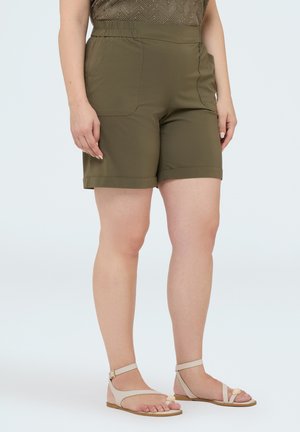 Kvinde iført olivengrønne knælange shorts med forlommer og beige flade sandaler med ankelremme mod en ensartet lys baggrund.