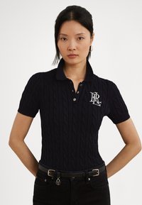 Tmavomodrá pletená polokošeľa s vrkočovým vzorom, krátkymi rukávmi a golierom. Doplnky zahŕňajú kontrastné vyšívané logo a gombíky.