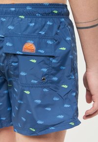 Shorts da bagno blu navy realizzati in un tessuto leggero e resistente all'acqua. Presentano una tasca posteriore con un logo arancione e piccoli motivi a foglia in blu e verde.