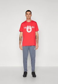 Rotes Baumwoll-T-Shirt mit "SWITZERLAND" Aufdruck in Weiß und Flammen, kombiniert mit grauer Jogginghose mit Seitenstreifen und schwarzen Schuhen.