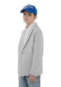 Blazer grigio con una texture liscia, chiusura a bottone singolo e due tasche, indossato sopra una camicia bianca. Abbinato a un berretto blu con testo bianco.