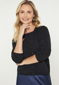 Musta neulecardigan, jossa on pyöreä kaula-aukko, pitkät hihat ja nappikiinnitys. Pehmeä rakenne, tyylikäs muotoilu, yhdistettynä siniseen satiinihameeseen.