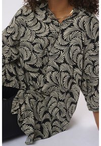 Chemise noire à manches longues avec des motifs abstraits de feuilles beiges portée par une personne aux cheveux bouclés et un collier argenté, assise sur un fond clair.