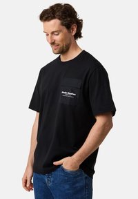 Musta puuvillapaita lyhyillä hihoilla, pyöreällä kaula-aukolla ja rintataskulla, jossa on valkoista tekstiä. Yhdistetty sinisiin farkkuihin.