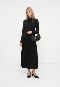 Melnas garās piedurknes blūze ar twist detaļu, melna plīsuma svārki, melnas pārvadājamo papēžu kurpes un melna brīva rokas soma. Visas drēbes ir vienotā krāsā.