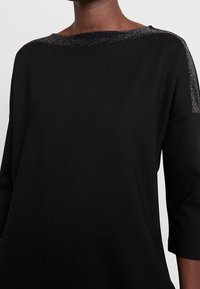 Haut noir avec un large décolleté orné d'un accent métallique, manches trois-quarts, et une texture lisse et douce. Design simple et élégant.