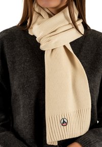 Écharpe beige à côtes fabriquée en matériau doux, ornée d'un patch logo de design circulaire avec des accents bleus et rouges. Portée avec un pull gris foncé.