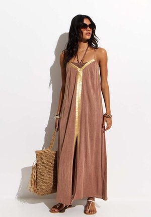 Femme portant une robe longue marron ornée de liserés dorés, des lunettes de soleil, des sandales et des bijoux, tenant un sac cabas en osier, debout devant un fond blanc.