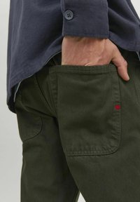 Olive green cotton pants with a textured finish, featuring a visible back pocket and red stitching detail. Paired with a navy button-up shirt.