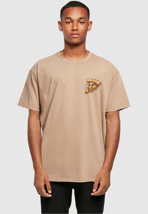 Young man wearing a beige t-shirt with a small dripping pepperoni pizza slice graphic on the left chest, paired with black pants.