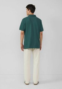 Dunkelgrünes Poloshirt mit Kragen und kurzen Ärmeln, kombiniert mit hellbeigen Hosen und braunen Schuhen, von hinten angezeigt.