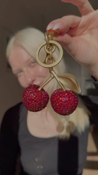 Rdeč obesek za ključe v obliki češnje iz smole z rahlo teksturirano površino, pritrjen na zlat kovinski obroček z listnim poudarkom.