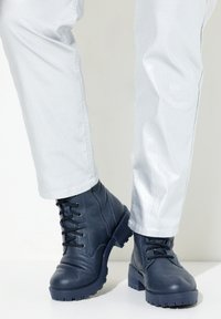 Botas de cuero navy hasta el tobillo con cordones, suela resistente y acabado texturizado, combinadas con pantalones metálicos plateados claros, mostrando un dobladillo recortado.